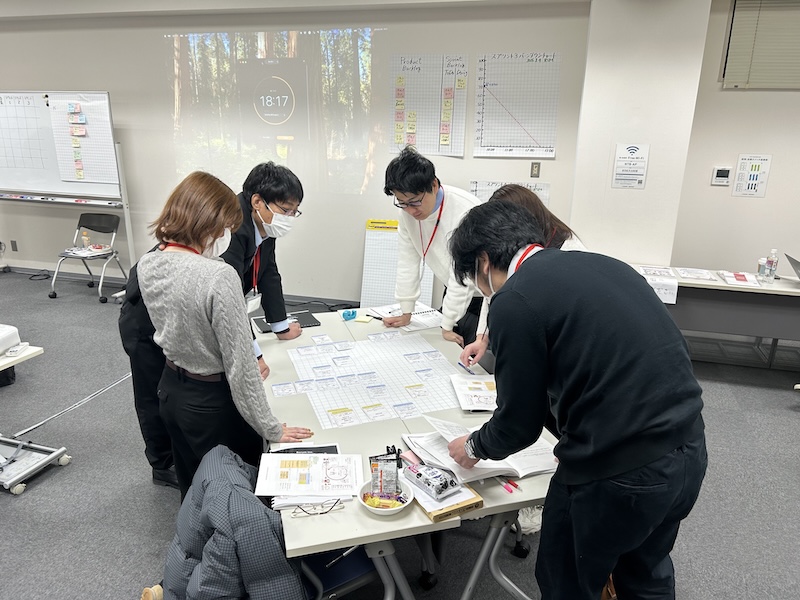 Five people wearing lanyards and masks stand around a table, examining papers and sticky-note cards on a planning board during a workshop in a conference room.