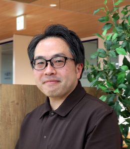 Portrait of a man with black hair and glasses wearing a brown polo, seated indoors near a wooden divider and green plant.