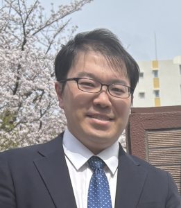 Portrait of a man in a suit and glasses outdoors, with cherry blossoms in the background.
